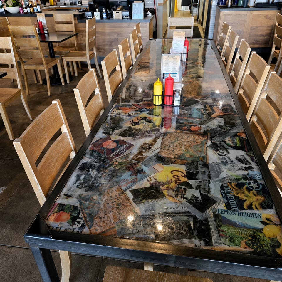 Long communal table with a collage top and wooden chairs in a bright, empty restaurant dining room.