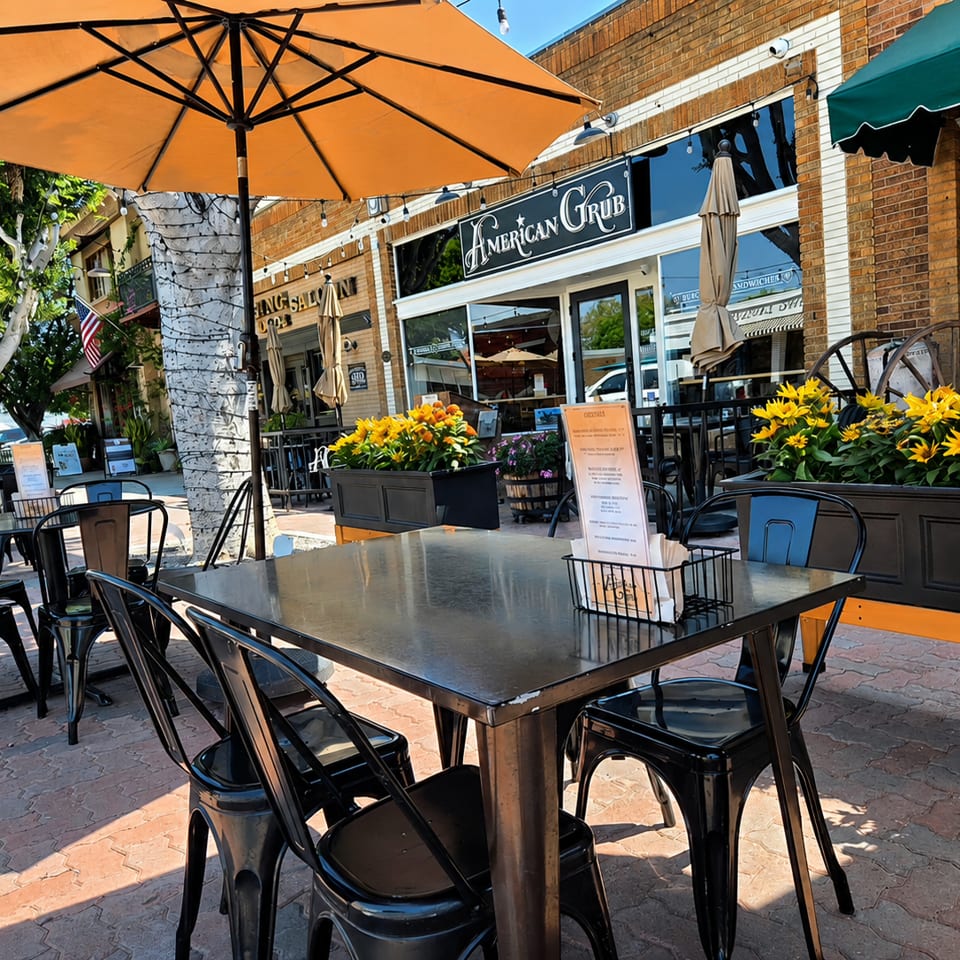 Outdoor patio seating at American Grub restaurant with black tables, chairs, and orange umbrellas.