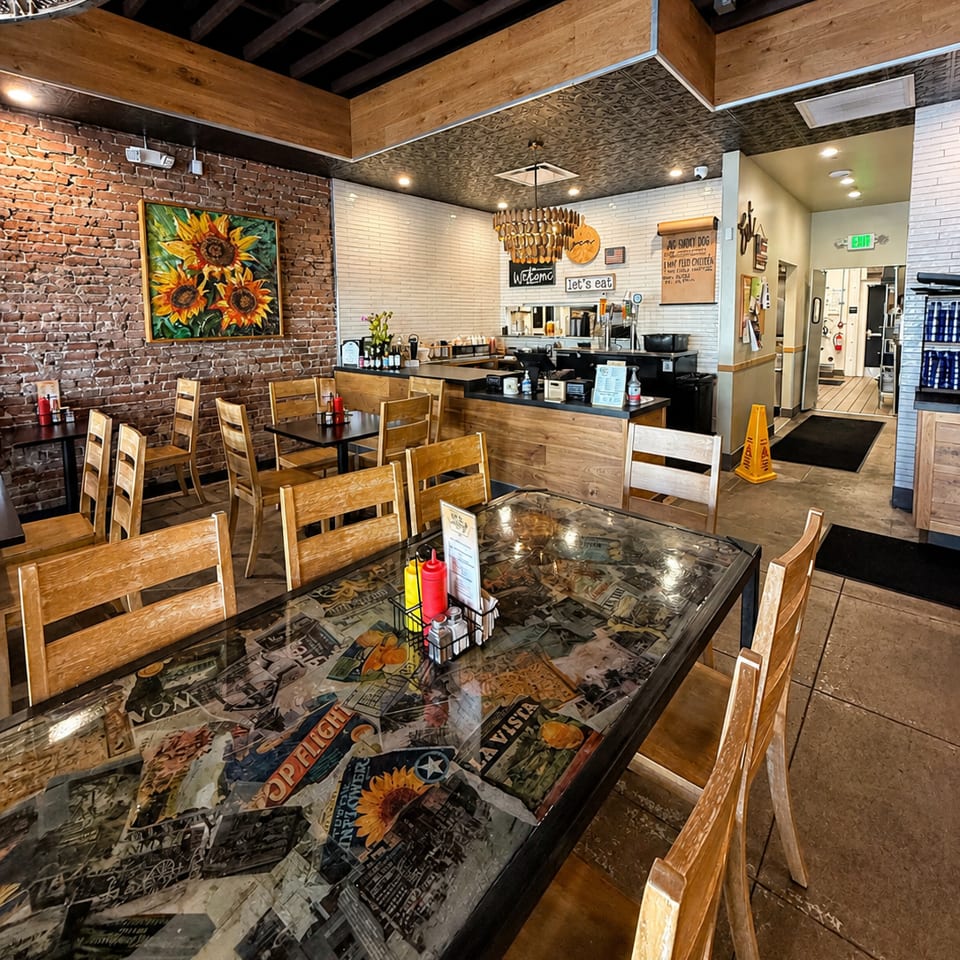 Restaurant interior with brick walls, wooden tables, chairs, and a counter area with a "Welcome" sign.