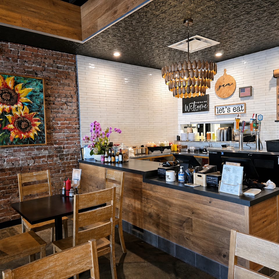Restaurant interior with a brick wall, sunflower painting, wooden tables, and a service counter.