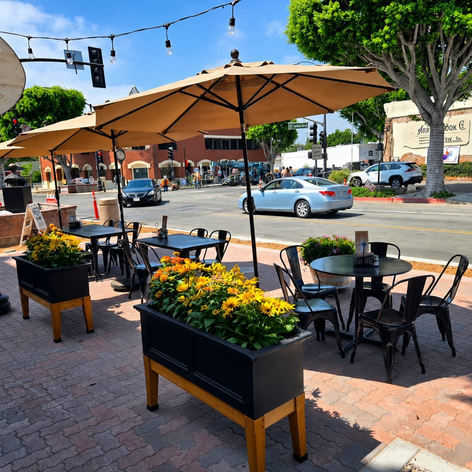 Outdoor patio dining area with black tables, chairs, and yellow flower planters under umbrellas on a sunny day.